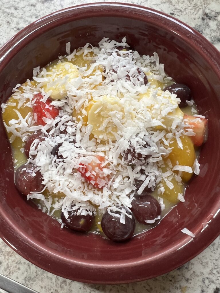 fruit salad with canned peaches, strawberries, red seedless grapes, sliced bananas topped with coconut flakes in a burgundy bowl